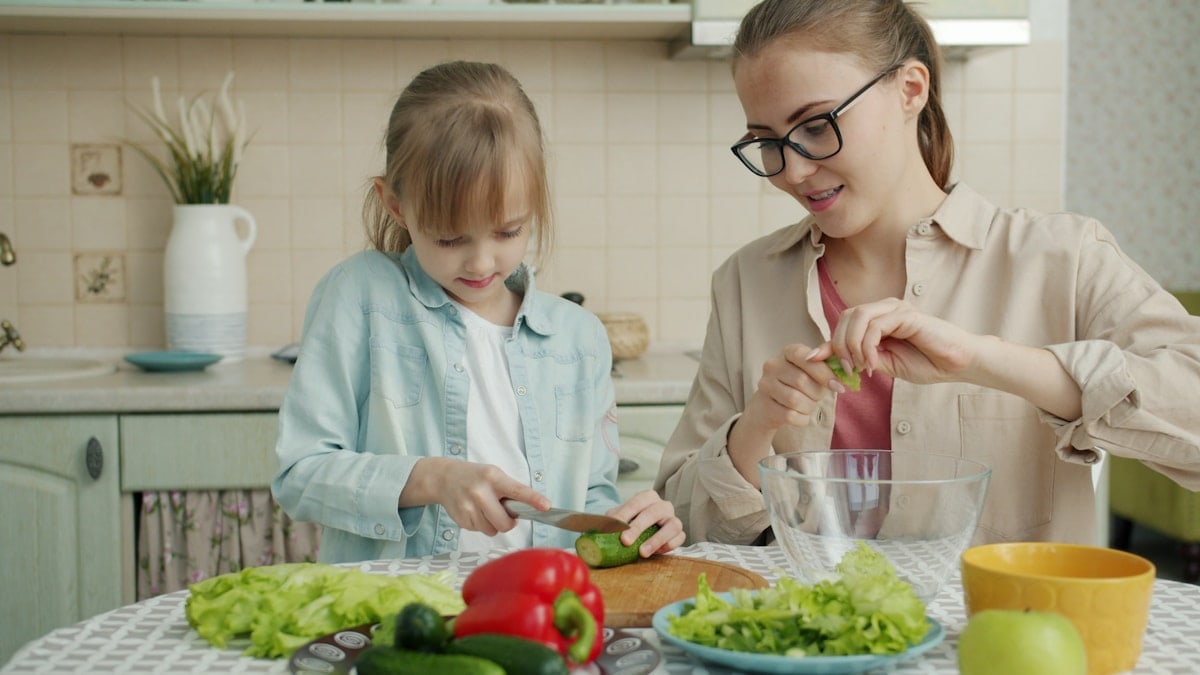 Adorable little girl is helping mother in kitchen cutting vegetables for salad and talking making dinner together. Healthy nutrition, lifestyle and people concept.